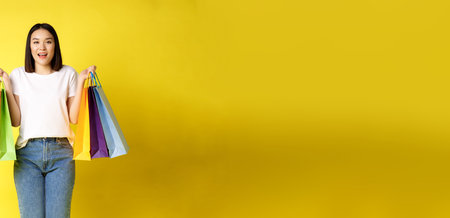Full size portrait of beautiful asian girl going shopping, holding paper bags from stores and smiling, standing in jeans and white t-shirt over yellow backgroundの写真素材