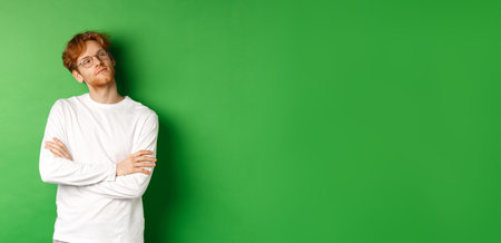 Confident young man with red hair, wearing glasses, looking smug at right side copy space, cross arms on chest self-assured, standing over green backgroundの写真素材