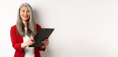 Elegant asian senior businesswoman working with documents, holding pen and clipboard, signing contract and smiling, standing over white backgroundの写真素材