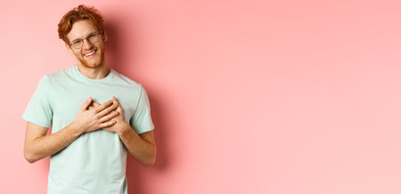 Attractive young man with ginger hair, holding hands on heart and smiling grateful, saying thank you, express gratitude, standing over pink backgroundの写真素材