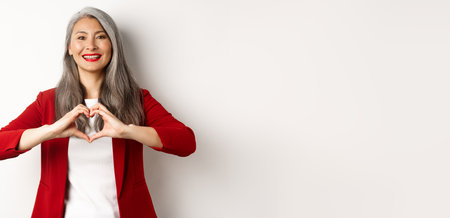 Beautiful asian mature woman in red blazer and makeup, showing heart sign and smiling, I love you gesture, standing over white backgroundの写真素材