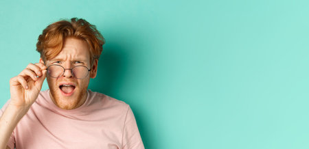 Close up portrait of redhead guy take-off glasses and looking confused at something strange, standing shocked over turquoise backgroundの写真素材