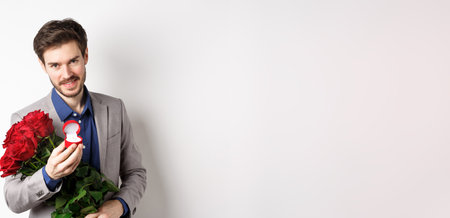 Romantic man with boquet of red roses asking to marry him, holding engagement ring and looking confident at camera, standing in suit over white backgroundの写真素材