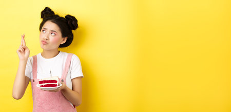 Holidays and celebration. Cute asian birthday girl making wish at party wish cake and candle, cross fingers and looking up, standing on yellow backgroundの写真素材