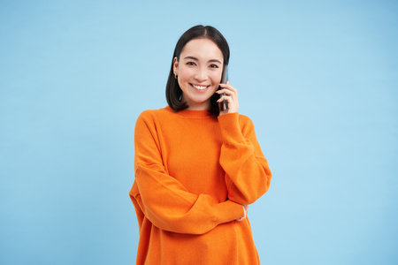 Portrait of young candid woman, asian girl talking on cellphone, holds mobile phone and speaks, stands over blue backgroundの写真素材