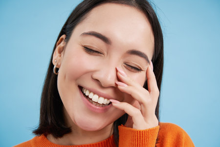 Close up portrait of candid happy woman, asian girl laughing and smiling, touches her clear skin, shows white teeth, blue backgroundの写真素材