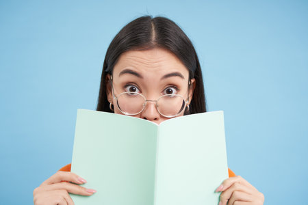 Portrait of cute japanese woman in glasses, peeking from notebook, covers face with her notes, smiles with eyes, blue backgroundの写真素材