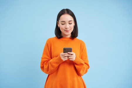 Cute asian girl looks at her cellphone and smiles, uses mobile application, social media app,...