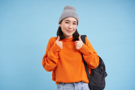 Cute korean woman shows thumbs up, approves smth good, likes and gives good feedback, recommends something, standing over blue backgroundの写真素材