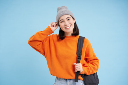 Stylish young asian woman in warm hat, walking with backpack, going somewhere with bag, standing over blue studio backgroundの写真素材
