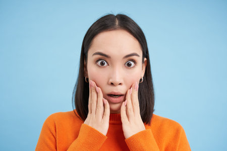 Close up portrait of asian girl with shocked face, looks at smth unbelievable, startled by news,...