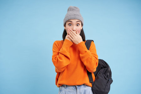 Portrait of korean girl in hat, covers her mouth with hands, looks shocked, amazed by gossips,...