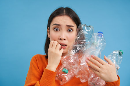 Surprised korean woman, holding plastic bottles, looking shocked at camera, concept of environment and recycling, blue backgroundの写真素材