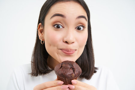 Close up of cute smiling asian woman, holding chocolate cupcake near mouth, having bite, enjoys eating pastry, white backgroundの写真素材