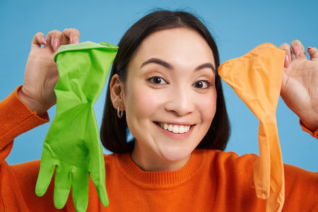 Close up portrait of smiling asian woman, showing two different latex gloves for cleaning, looking enthusiastic, doing house errands, blue backgroundの写真素材
