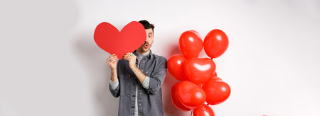 Cute young man hiding face behind big Valentine heart card, secret admirer looking at camera with amazement, saying wow, standing near romantic balloons on white backgroundの写真素材