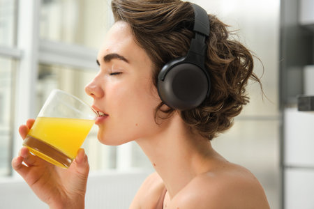 Smiling sportswoman, fitness girl does workout at home, wears headphones, listens music and drinks orange juice after training sessionの写真素材