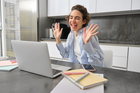 Beautiful girl waves hands at laptop camera, says hello to coworkers, connects to work meeting...