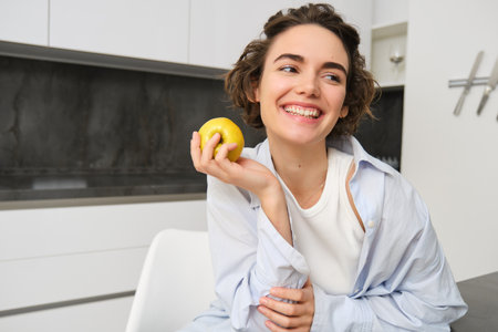 Image of brunette woman laughing, smiling while eating an apple in kitchen, sitting at home, having a snackの写真素材