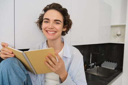 Portrait of smiling young woman reading journal, enjoys comfort at home, holding notebook, looking happy, relaxing indoorsの写真素材