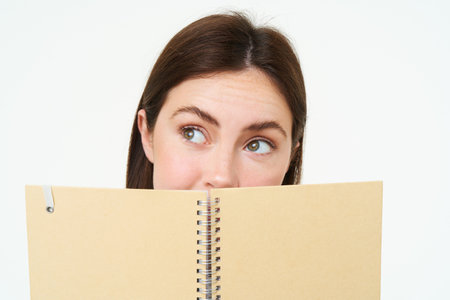 Young woman holds notebook daily planner next to her face, writing down homework, making notes, looking thoughtful, standing over white backgroundの写真素材