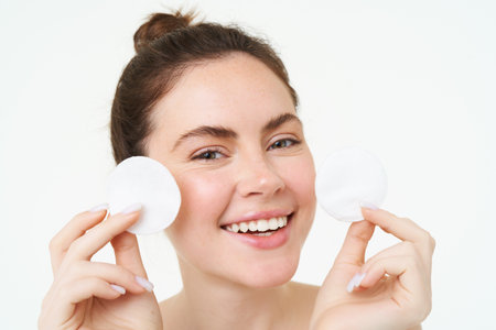 Beauty woman with clear glowing face, showing cotton cosmetic pads for makeup removal, cleansing her facial skin, standing over white backgroundの写真素材