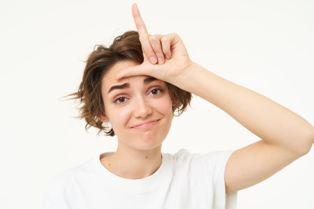 Close up of woman shows loser gesture, l letter on forehead and smiling, mocking, makes fun of someone, standing over white backgroundの写真素材