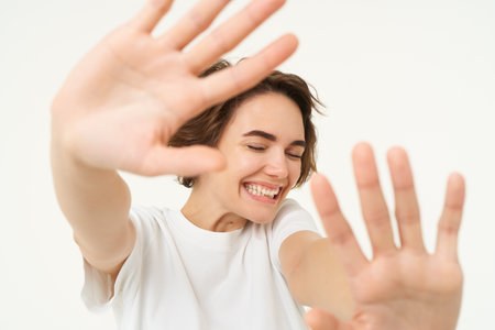 Close up of happy brunette woman extending her hands, stretching, covering herself from camera, laughing and smiling, standing over white backgroundの写真素材