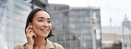 Vertical shot of smiling asian woman in wireless headphones, enjoys listening to music in earphones, holds mobile phoneの写真素材