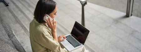 Portrait of asian girl, student sits on stairs with laptop, talks on mobile phone. Young woman makes a telephone call while working on computerの写真素材