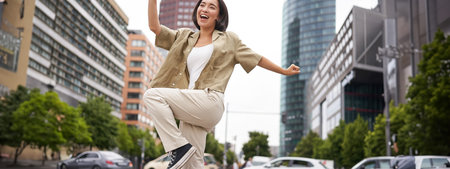 Portrait of dancing asian woman, raising hands up and feeling happy, posing in city, triumphing,...