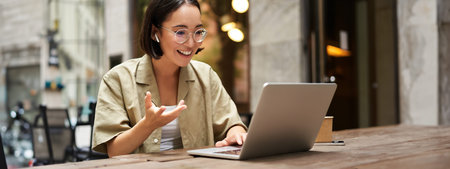 Online meeting. Young urban girl, asian woman talking with laptop, video chat, gesturing, sitting in...