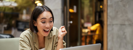 Vertical shot of happy girl talking on video call, looks at laptop, having online meeting, sitting in outdoor cafeの写真素材