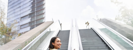 Young asian girl going up on an escalator, holding smartphone, smiling while walking in city