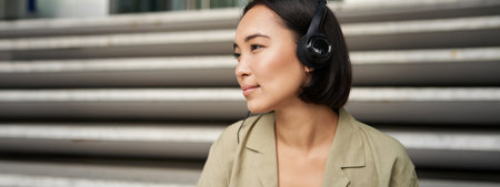 Young asian woman listening to music in headphones, sitting outside on street, smilingの写真素材