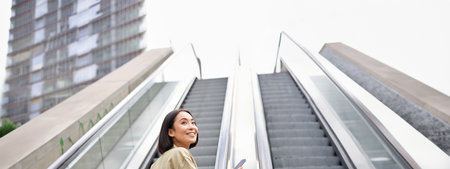Young asian girl going up on an escalator, holding smartphone, smiling while walking in cityの写真素材