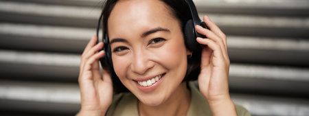 Close up portrait of smiling asian girl in headphones, listens to music outdoors, looking happy.の写真素材