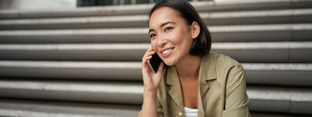 Portrait of beautiful asian girl talks on mobile phone, sits on street stairs. Woman with smartphone smiling, making a callの写真素材