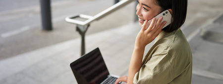 Portrait of asian girl, student sits on stairs with laptop, talks on mobile phone. Young woman makes...