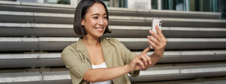 Portrait of asian beautiful girl takes selfie on smartphone. Young korean woman sits on stairs outdoors and makes photos on mobile phoneの写真素材