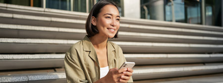 People and technology. Smiling beautiful asian woman sitting on stairs in city, holding mobile phone. Girl with smartphone rests outsideの写真素材