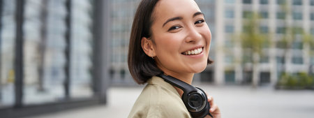 Portrait of happy asian woman in headphones, enjoys music while walking in city, smiling and laughingの写真素材