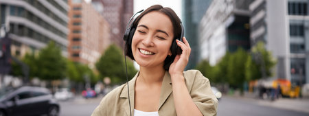 Portrait of smiling asian woman in headphones, standing in city centre on street, looking happy, listening to musicの写真素材