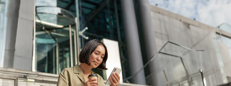 Beautiful smiling asian girl, drinking coffee, using mobile phone and sits on stairs outside. Young...