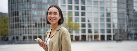 Vertical shot of young asian digital nomad, girl talks on mobile phone and walks on street with laptop. Young woman remote worker going to coworking spaceの写真素材