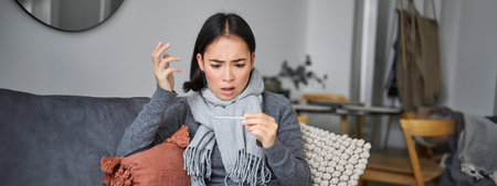 Shocked woman looks concerned at her thermometer, catching cold, has fever, sitting on sick leave at...
