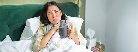 Portrait of couching asian girl in bed, feeling sick, catching cold and staying at home, looking unhealthy, drinking hot teaの写真素材