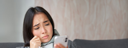 Ill korean woman sitting at home, sneezing, caught cold, reading news on mobile phone, freezing at her home because of heating problemsの写真素材