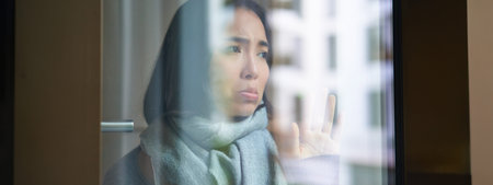 Portrait of sad ill korean woman looking out of the window, staying at home due to coronavirus,...