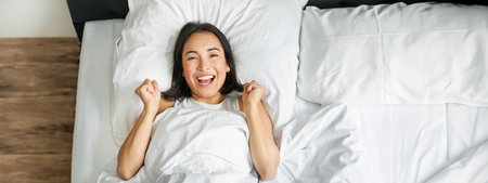 Top view of enthusiastic asian girl cheering, lying in her bed and shouting with joy and excitement, triumphing and smiling, enjoy staying in her bedroomの写真素材